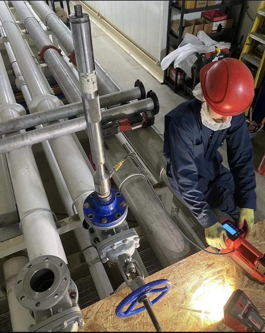 IMW technician in hard hat and protective gear working a hot tap among dense overhead plant piping