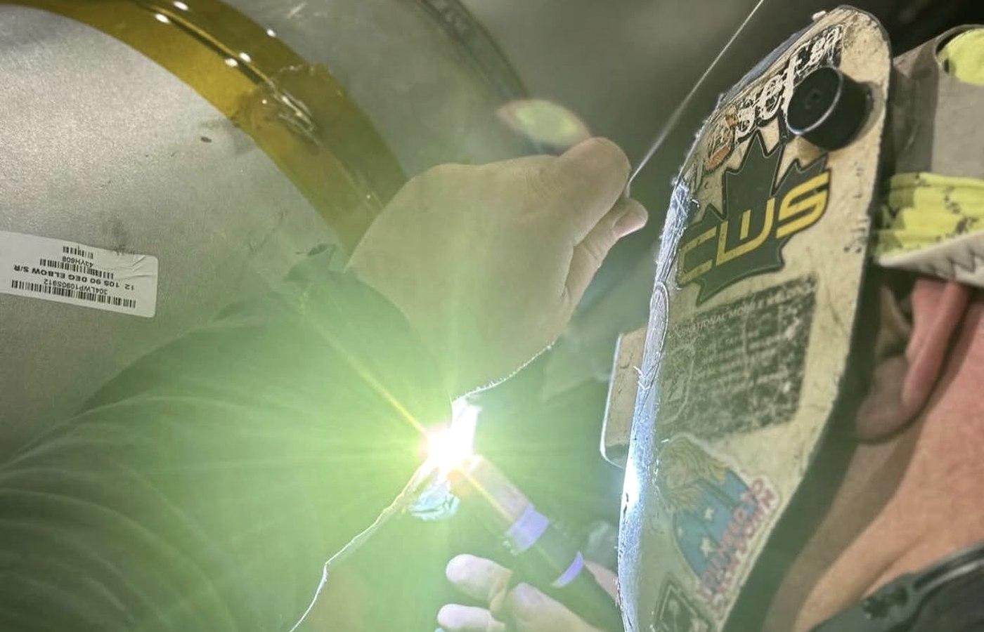 Hand-fed GTAW pass on a stainless steel elbow, welder in a CWB-stickered hood